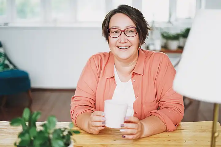Eine Frau mittleren Alters sitzt entspannt mit einer Tasse Kaffee in der Hand an einem Tisch und lächelt selbstbewusst und zuversichtlich in die Kamera.